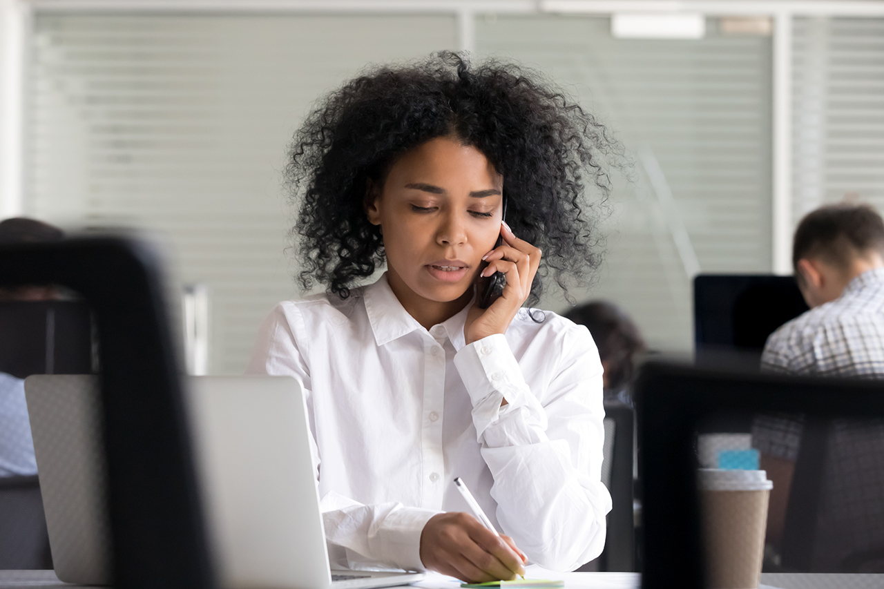 Focused African American woman making notes on sticker, clarifies information, talking on phone, registers complaint from client or customer, female employee consulting by mobile device at workplace