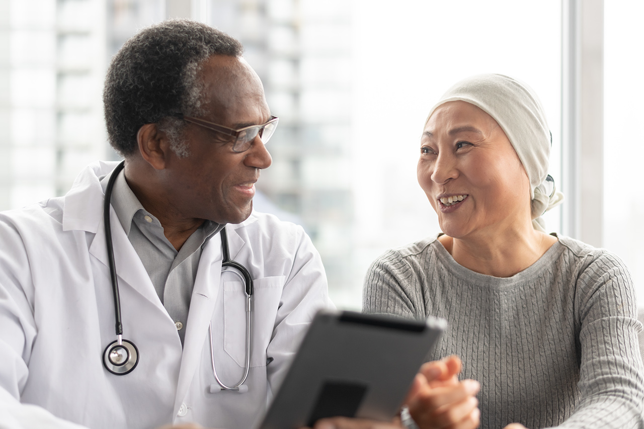 A Korean woman with cancer is meeting with her African American male doctor. The doctor is showing her test results on an electronic wireless tablet. The doctor smiles as he gives good news regarding the patient's treatment.