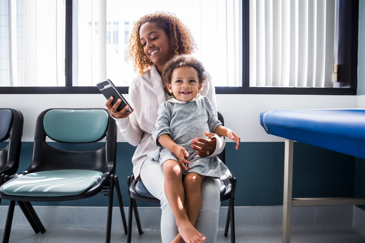 Mother using smart phone while toddler sitting on lap at doctors office