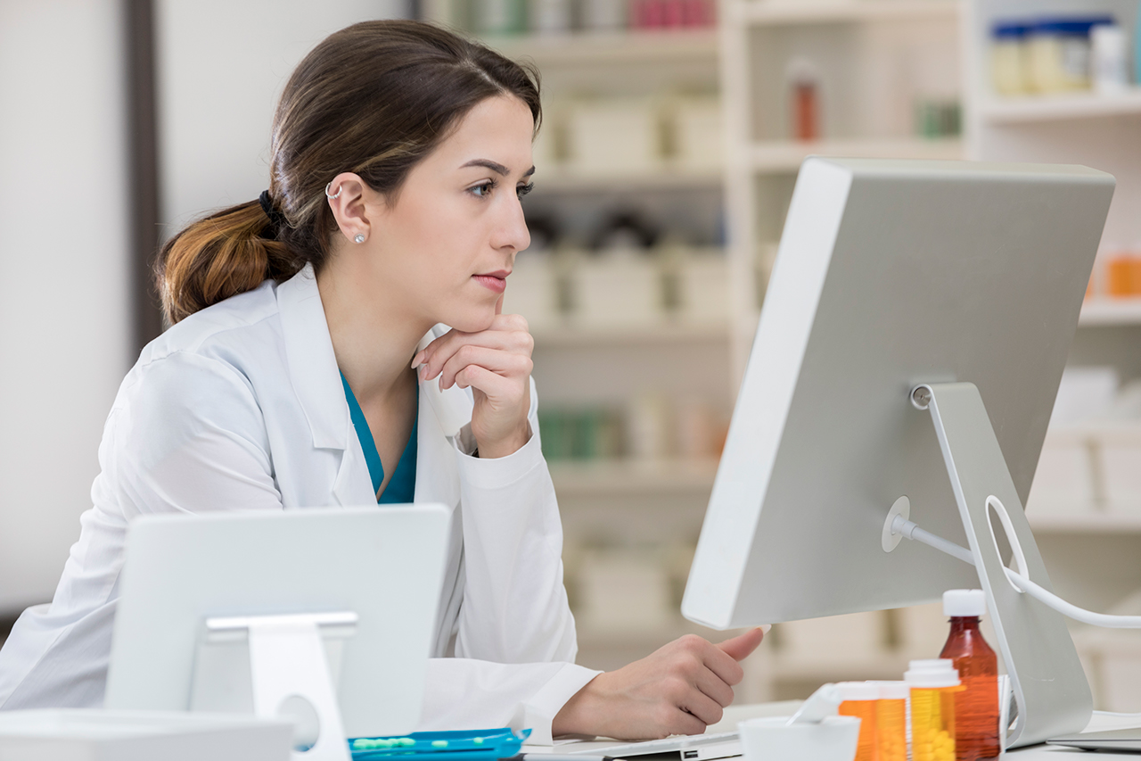 A serious Caucasian pharmacist leans on her hand at a table as she studies the latest incoming prescriptions on a computer screen.  There is pharmaceutical equipment in the foreground and shelves in the background.