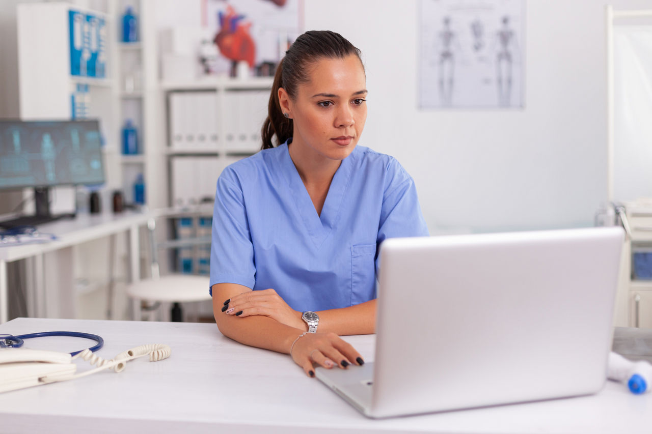 Medical nurse working on laptop in hospital office wearing blue uniform. Health care practitioner sitting at desk using computer in modern clinic looking at monitor, medicine.
